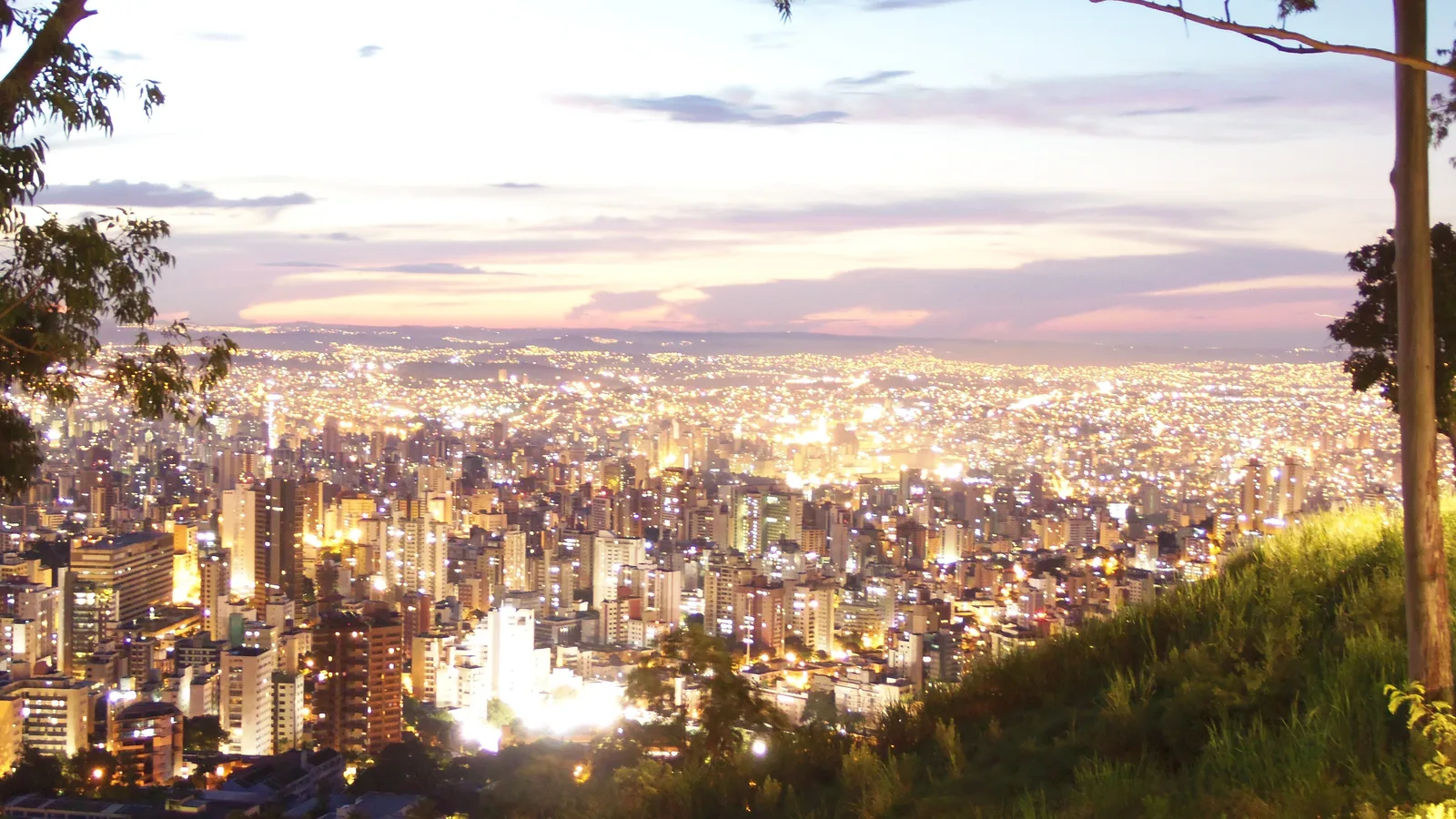 Belo Horizonte at dusk from a hillside: the city lights coming on across the whole valley under a pink and lilac sky, a dark grassy slope in the foreground
