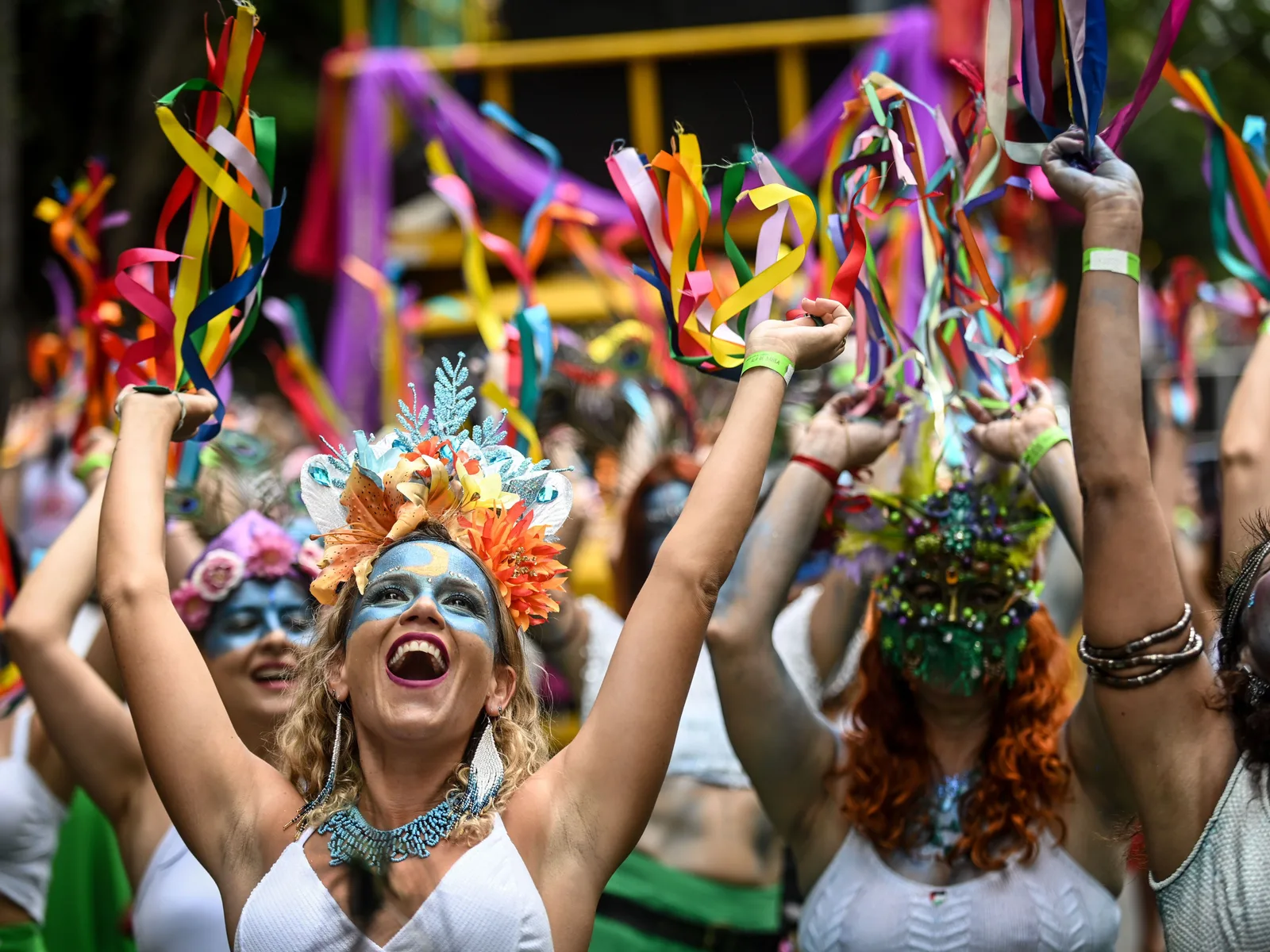 Revelers at a Belo Horizonte Carnaval bloco with painted faces and flower crowns, arms raised and hands full of colored ribbons, laughing in a packed crowd