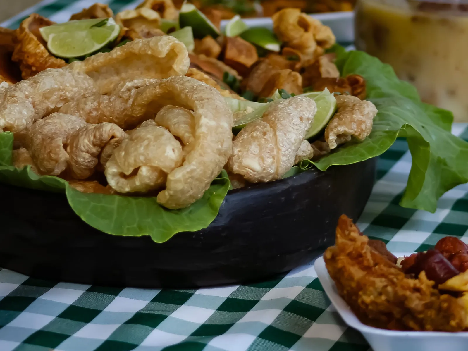 A black dish piled with golden pork crackling on lettuce leaves with wedges of lime, on a green-and-white checked tablecloth beside another plate of fried snacks