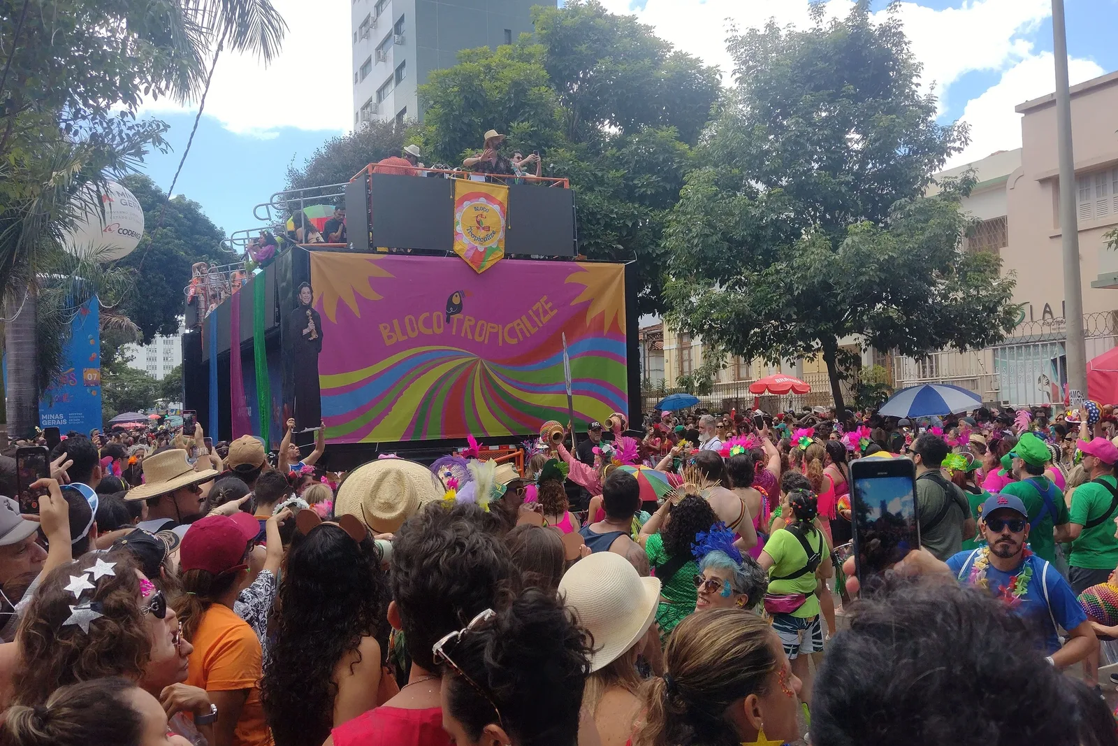 A dense street crowd in costume and glitter at a Belo Horizonte Carnaval bloco, a sound truck painted in bright colors behind them