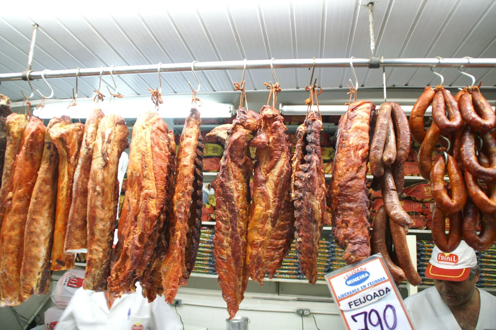 Cured sausages and slabs of dried pork hanging in a row over a butcher’s counter at the Mercado Central, a printed feijoada price card and a vendor in a cap below them