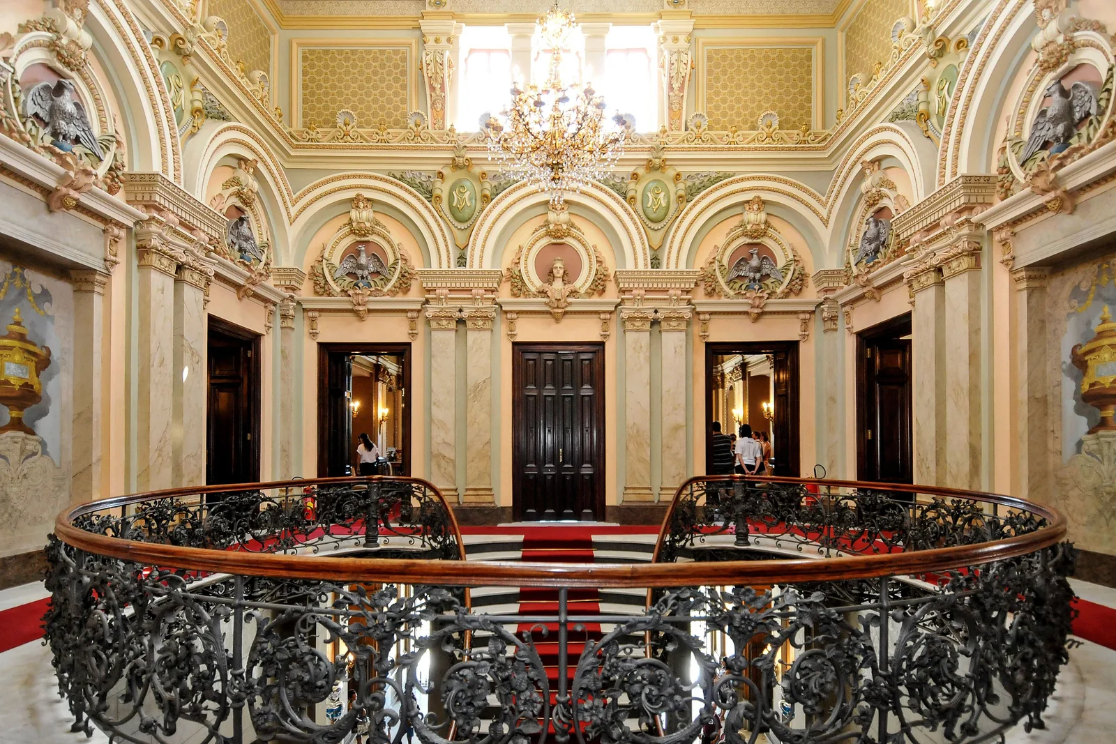 The gilded entrance hall of the Palácio da Liberdade from the upper floor: a wrought-iron balustrade around the stairwell, painted walls and a crystal chandelier