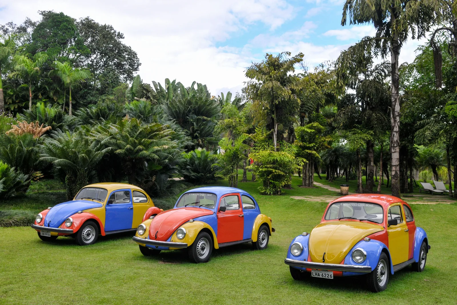Jarbas Lopes’s Troca-troca at Inhotim: three Volkswagen Beetles painted in patchwork blocks of red, yellow and blue parked on a lawn among palms and tropical trees