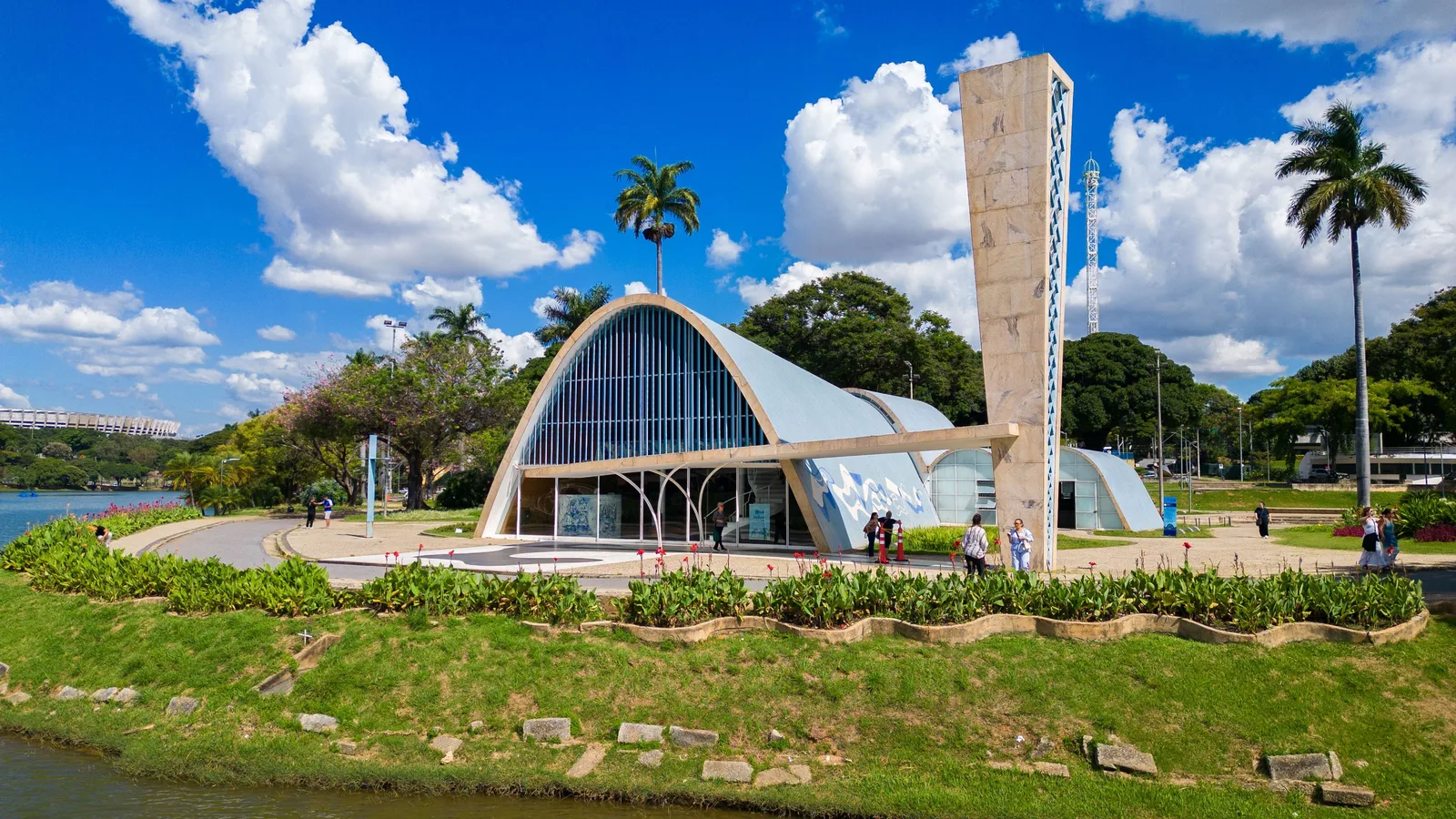 The São Francisco de Assis church at Pampulha seen across the water: a pale blue parabolic concrete vault with a glass front, its tall freestanding bell tower beside it, lawns and palms around it under a bright sky full of white cloud