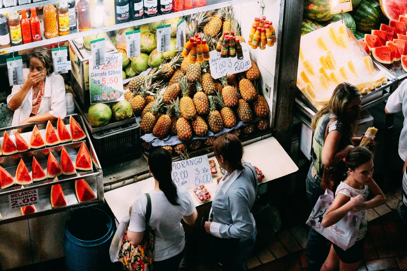 Looking down into a lane of the Mercado Central: a fruit stall stacked with pineapples and watermelon, shoppers in the aisle below