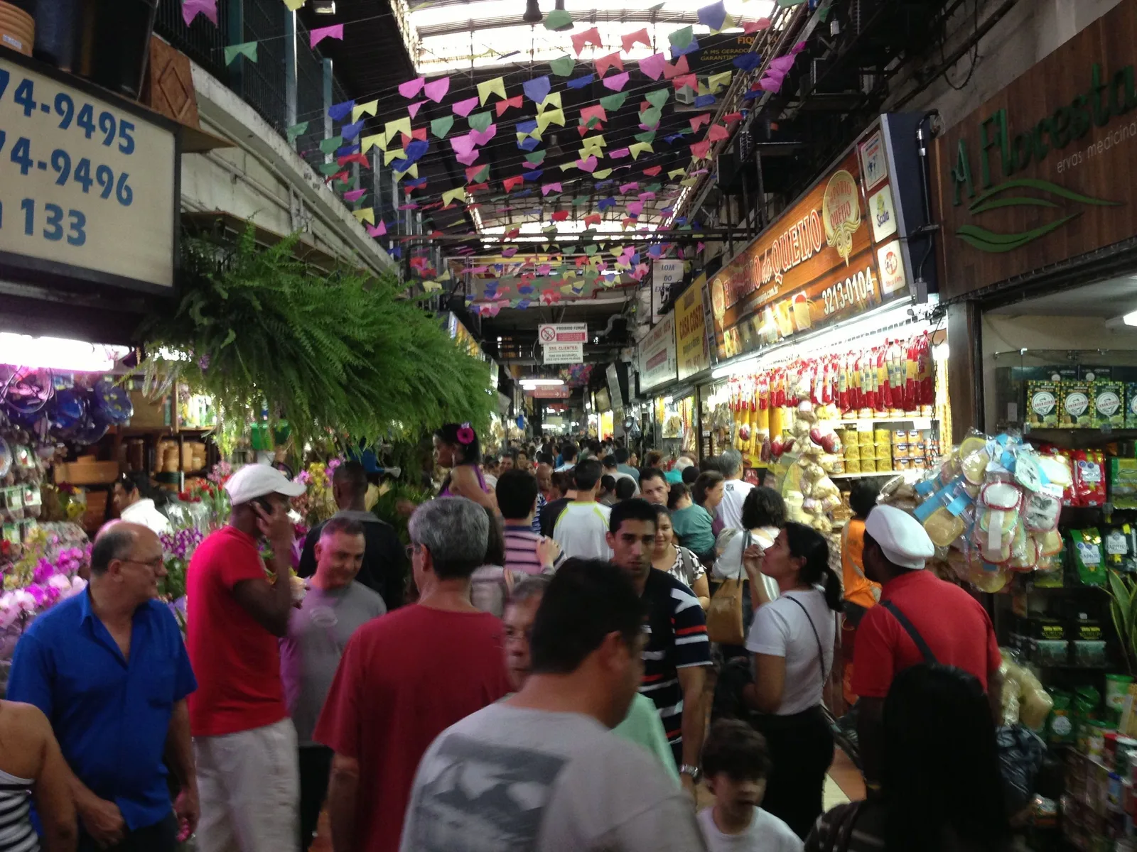 A crowded lane inside the Mercado Central: shoppers packed between stalls of flowers, plants and groceries, paper bunting strung overhead under the market roof