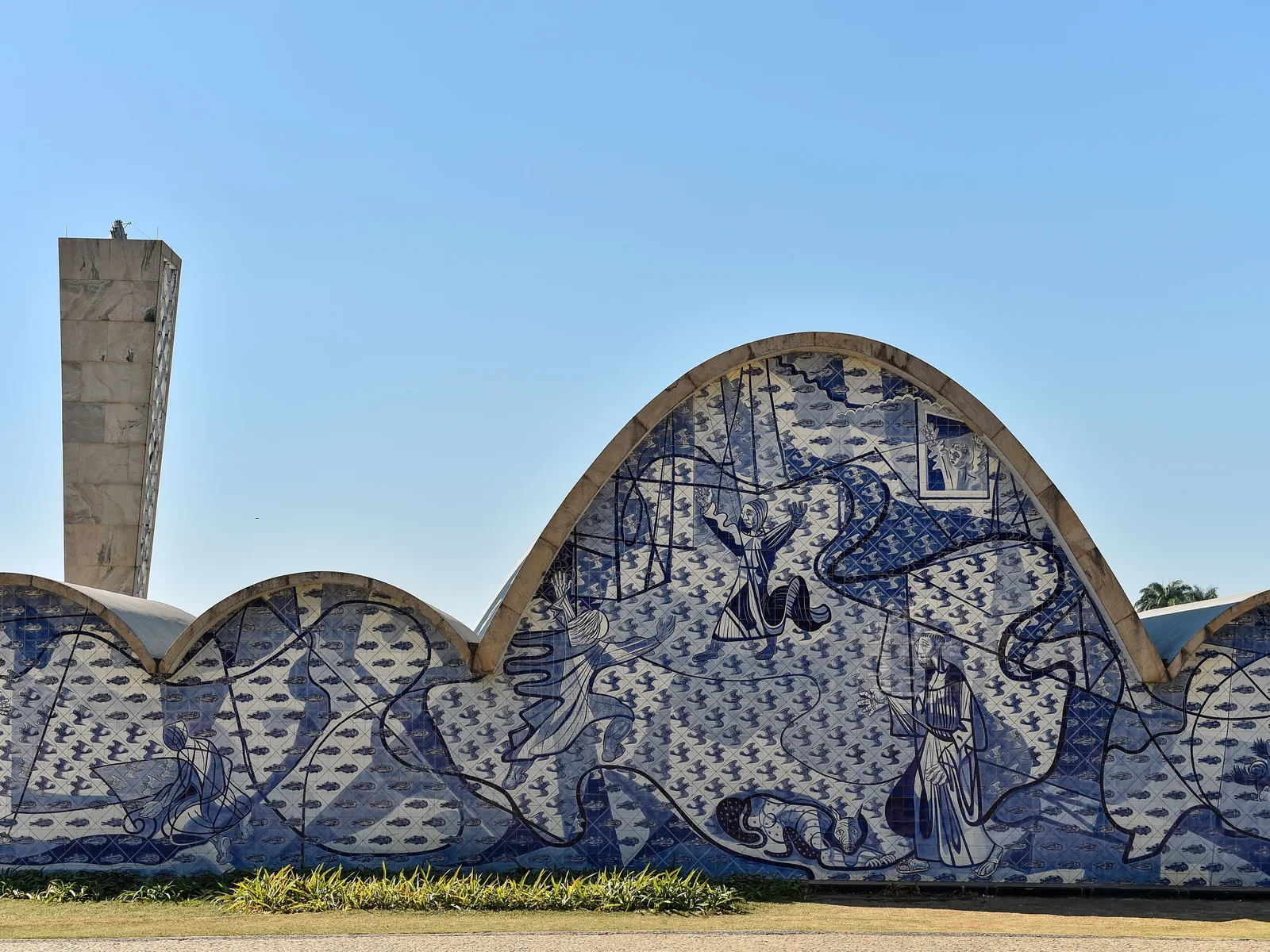 The back of the Pampulha church: a row of low concrete vaults rising to one tall arch, the whole wall covered in Cândido Portinari’s blue-and-white tile panel of figures and birds, the slim bell tower standing against the blue sky at the left