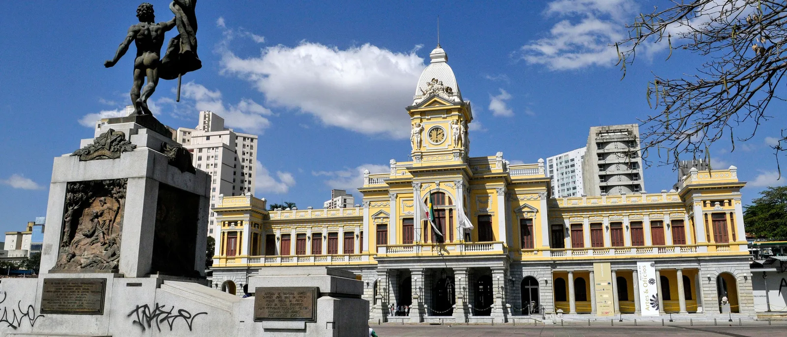 The Praça da Estação in central Belo Horizonte: the cream and yellow train station building with its clock tower behind a bronze statue on a tall plinth, under a blue sky