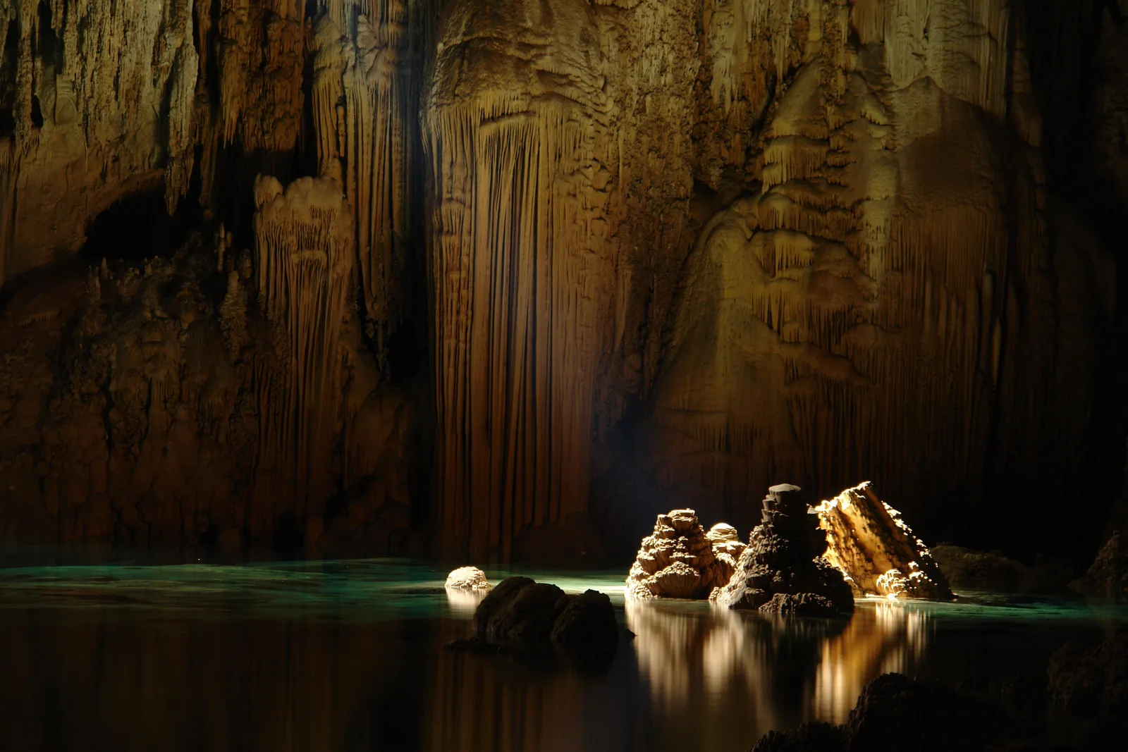 Warm-lit cone-shaped rock formations standing out of a still dark lake inside a cavern, reflected in the water, with a stalactite wall rising behind them