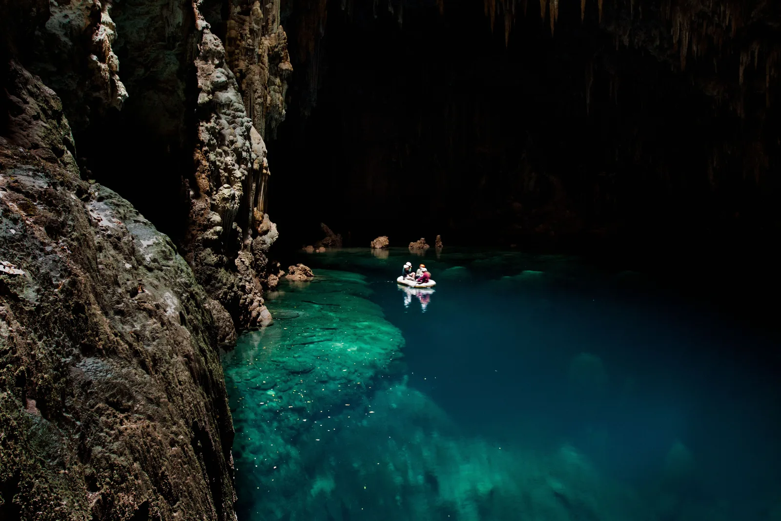 Looking down into the Abismo Anhumas at a turquoise lake at the bottom of a rock shaft, with two people on an inflatable raft floating on it