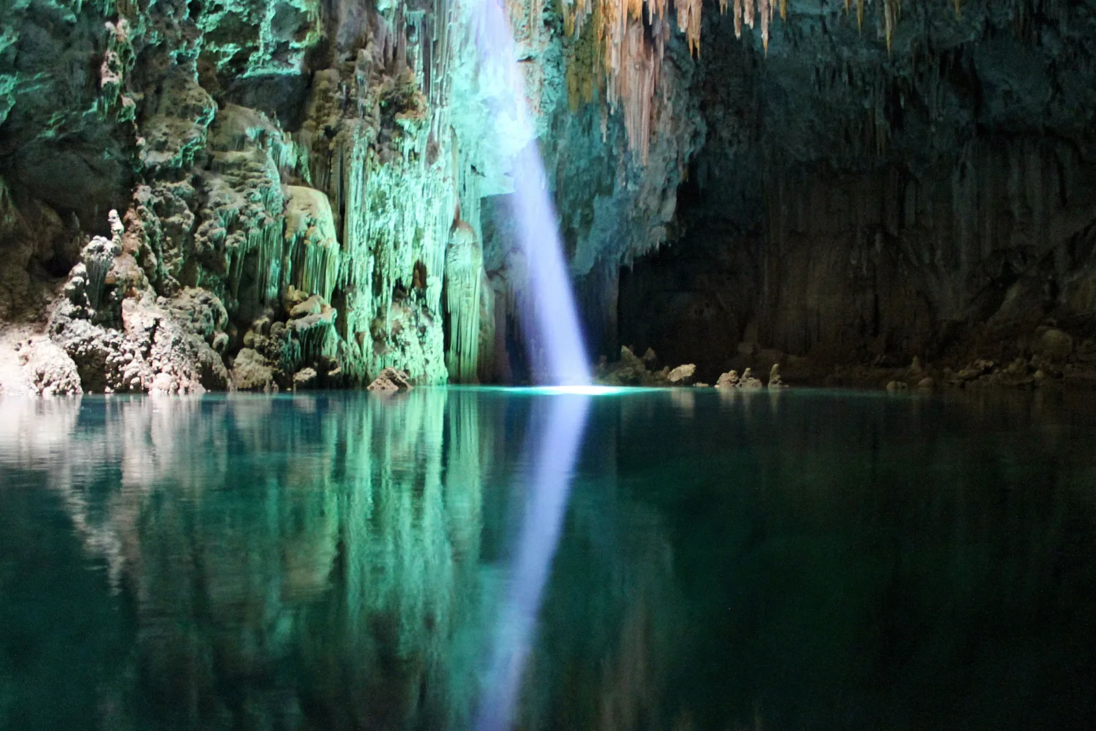 A shaft of daylight falling through the roof of the Abismo Anhumas onto the green lake at the bottom, stalactites hanging around it in the dark