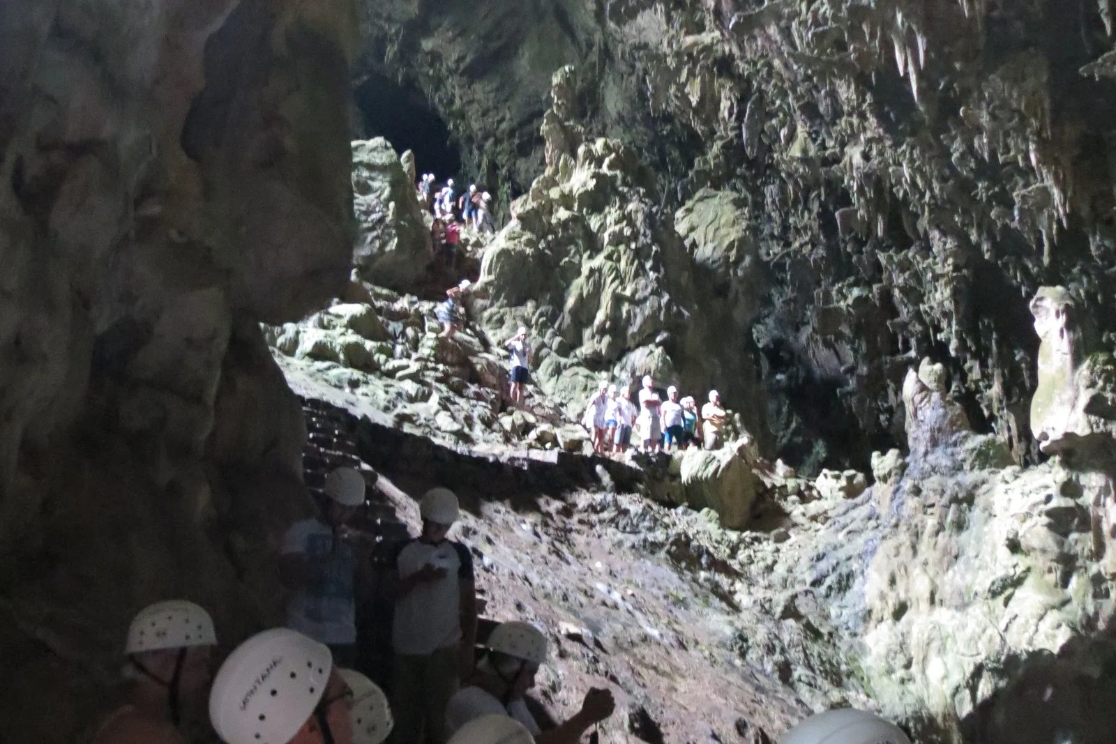 A group of visitors in white helmets picking their way down the pale rocky floor of a large cave chamber, with the cave walls rising steeply around them