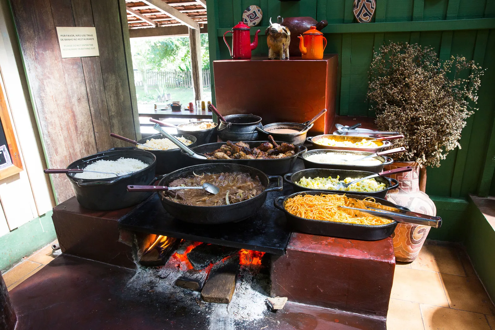 A wood-fired farm kitchen with a row of open pans of stews, rice and beans over a fire, jars and dried plants on a shelf above and a green wooden wall behind
