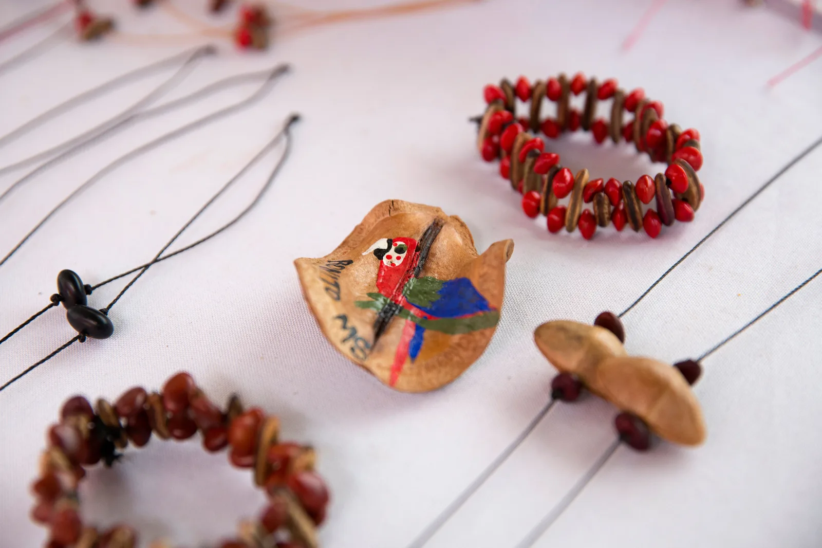 Handmade necklaces of red seeds and a painted macaw on a seed pod laid out on a white cloth at a craft stall in Bonito
