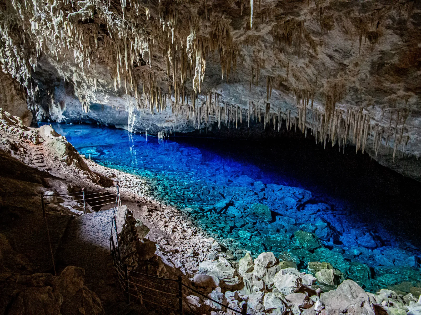 The interior of the Gruta do Lago Azul: a ceiling of pale stalactites over a brilliant blue underground lake, with a metal staircase running down the rock on the left