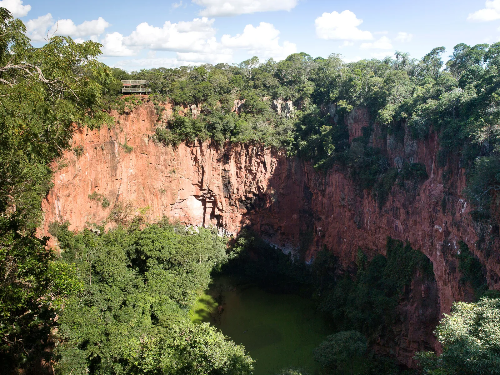 The Buraco das Araras seen from the rim: a vast circular pit with red-orange sandstone walls, dense green forest on the floor and a green pool at the bottom, with a steel viewing platform on the far rim