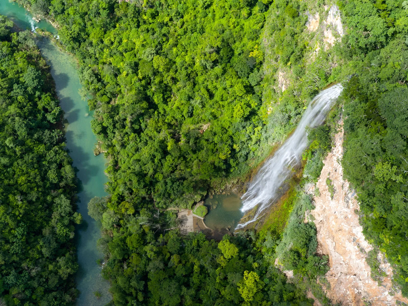 An aerial view of a tall thin waterfall dropping down a pale cliff into a forested canyon, with a turquoise river running along the canyon floor below it