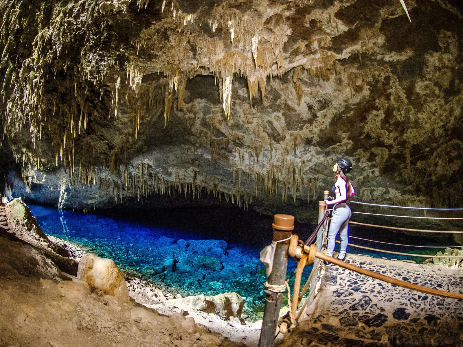 A visitor in a helmet standing at a rope handrail inside the Gruta do Lago Azul, the deep blue underground lake below, with a ceiling of pale stalactites lit from the entrance behind them