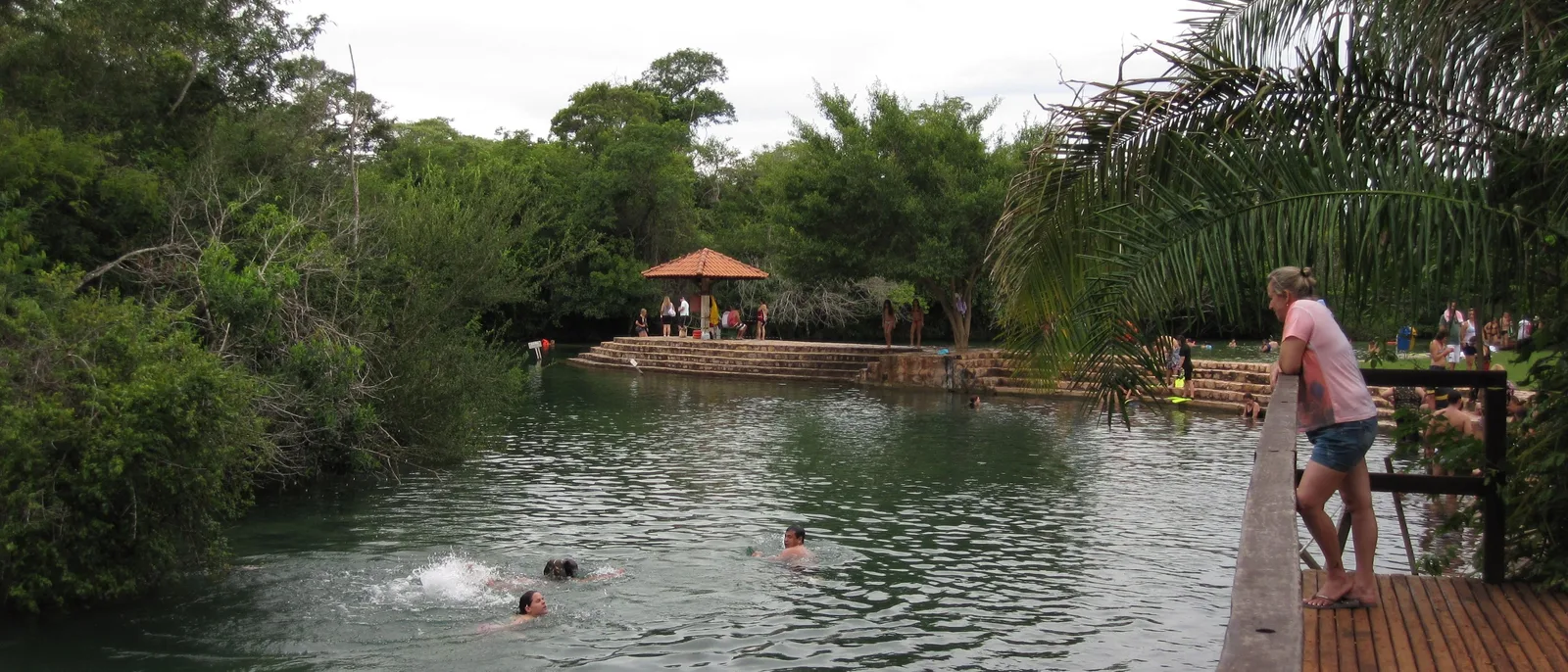A calm stretch of the Rio Formoso with a wooden deck and a thatched shelter on the bank, and people swimming in the clear water in front of them