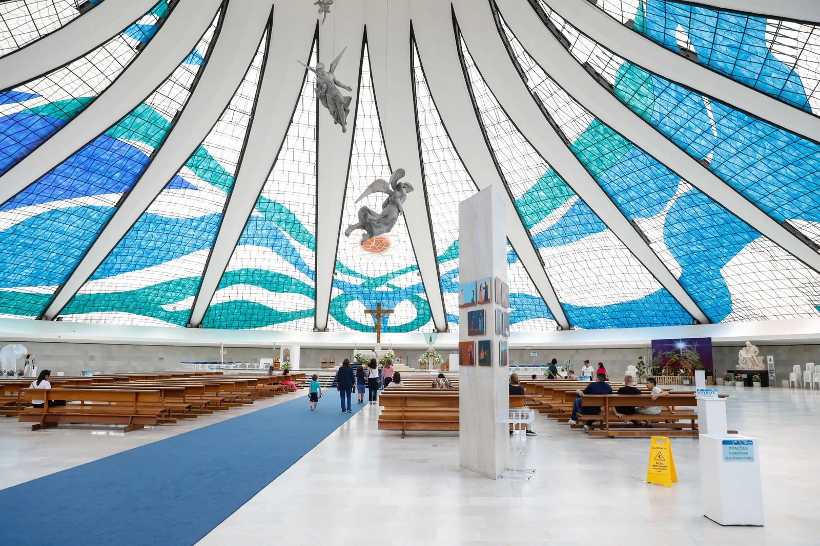 The nave of the Cathedral of Brasília: white concrete ribs sweeping up to the center, Marianne Peretti’s blue, green and white stained glass between them, an angel by Alfredo Ceschiatti hanging in the light, and rows of wooden pews below