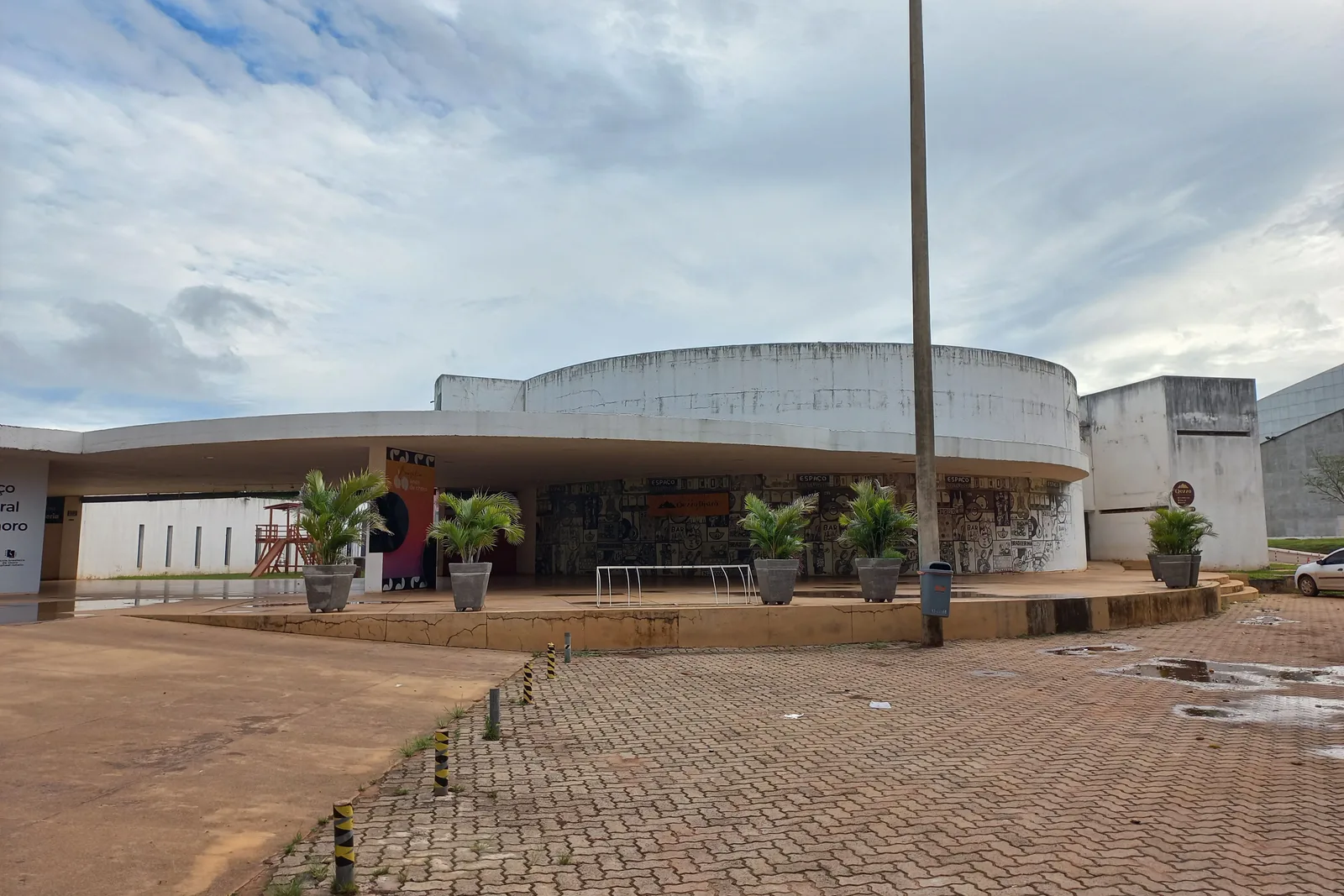 The Clube do Choro building in Brasília: a low pavilion with a curved white roof behind a paved forecourt and a tall lamp post, under a cloudy sky