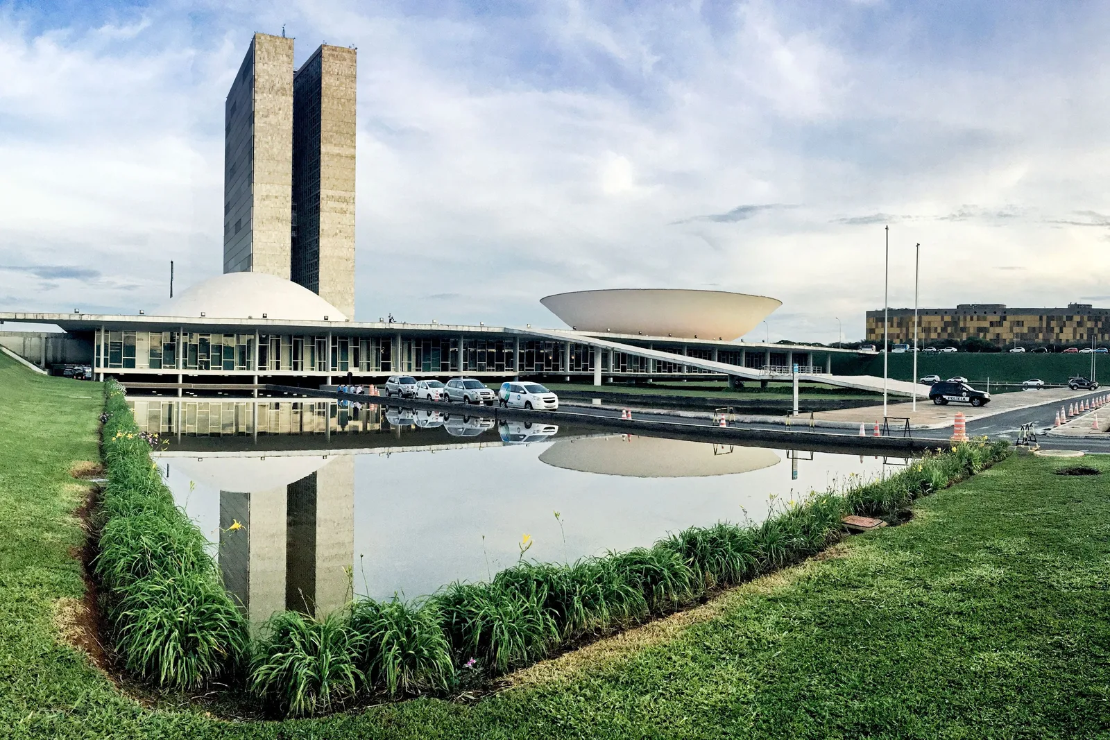 The National Congress from across a reflecting pool lined with planted beds: the twin towers between the upturned bowl and the dome, green lawns sloping down to the water and a cloudy sky