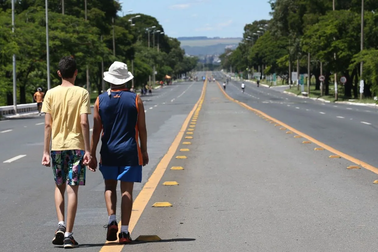 The Eixão closed to cars on a sunny day: two people walking away down the empty lanes beside the yellow center line, runners and cyclists far ahead, and tall trees and lamp posts lining both sides