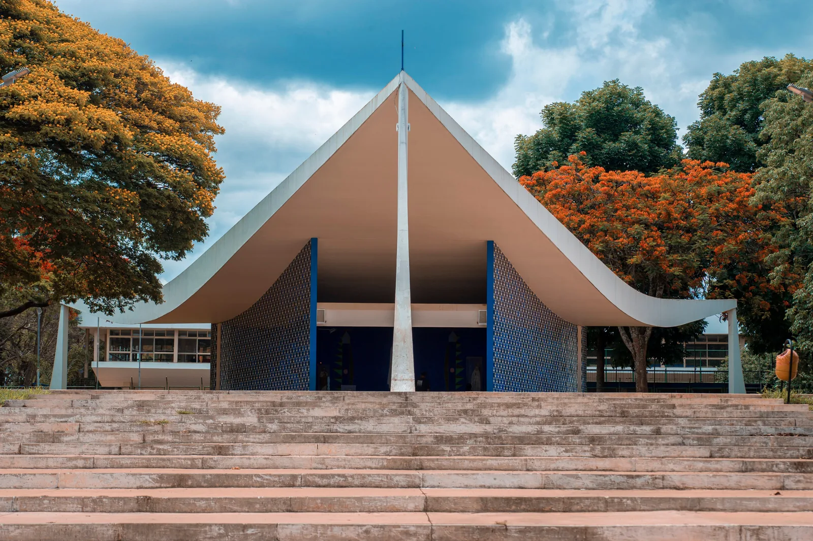 The Igrejinha in Asa Sul: a white triangular concrete roof folding down to three points, the blue tiled walls of the chapel beneath it, a golden-flowering tree on the left and a red flamboyant on the right, and steps in front