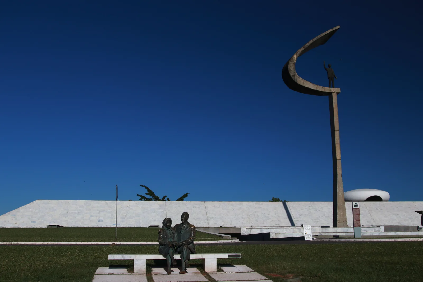The Memorial JK on a bright day: a low white building on a lawn and, at the right, the tall slim pylon with its curved crook holding the statue of Juscelino Kubitschek, bronze figures of JK and his wife seated on a bench in the foreground