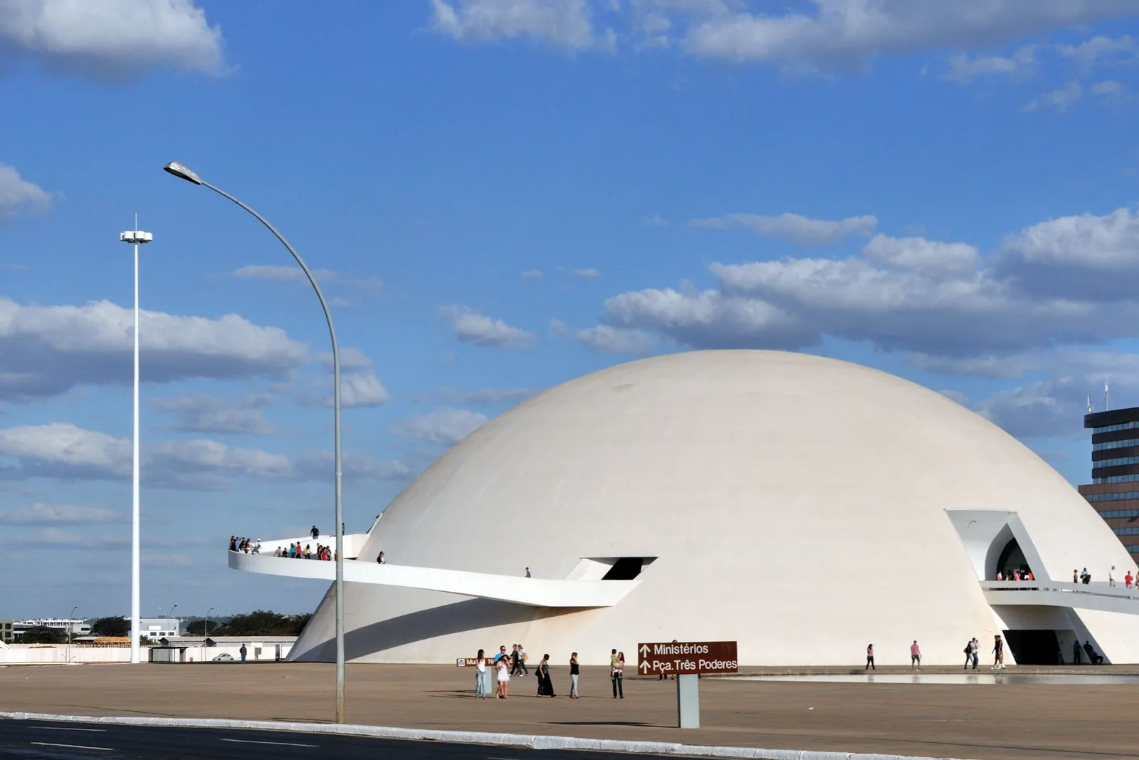 The white dome of the Museu Nacional with a curved ramp wrapping out of its side, visitors on the paved forecourt and a sign pointing to the ministries and the Praça dos Três Poderes, under a blue sky