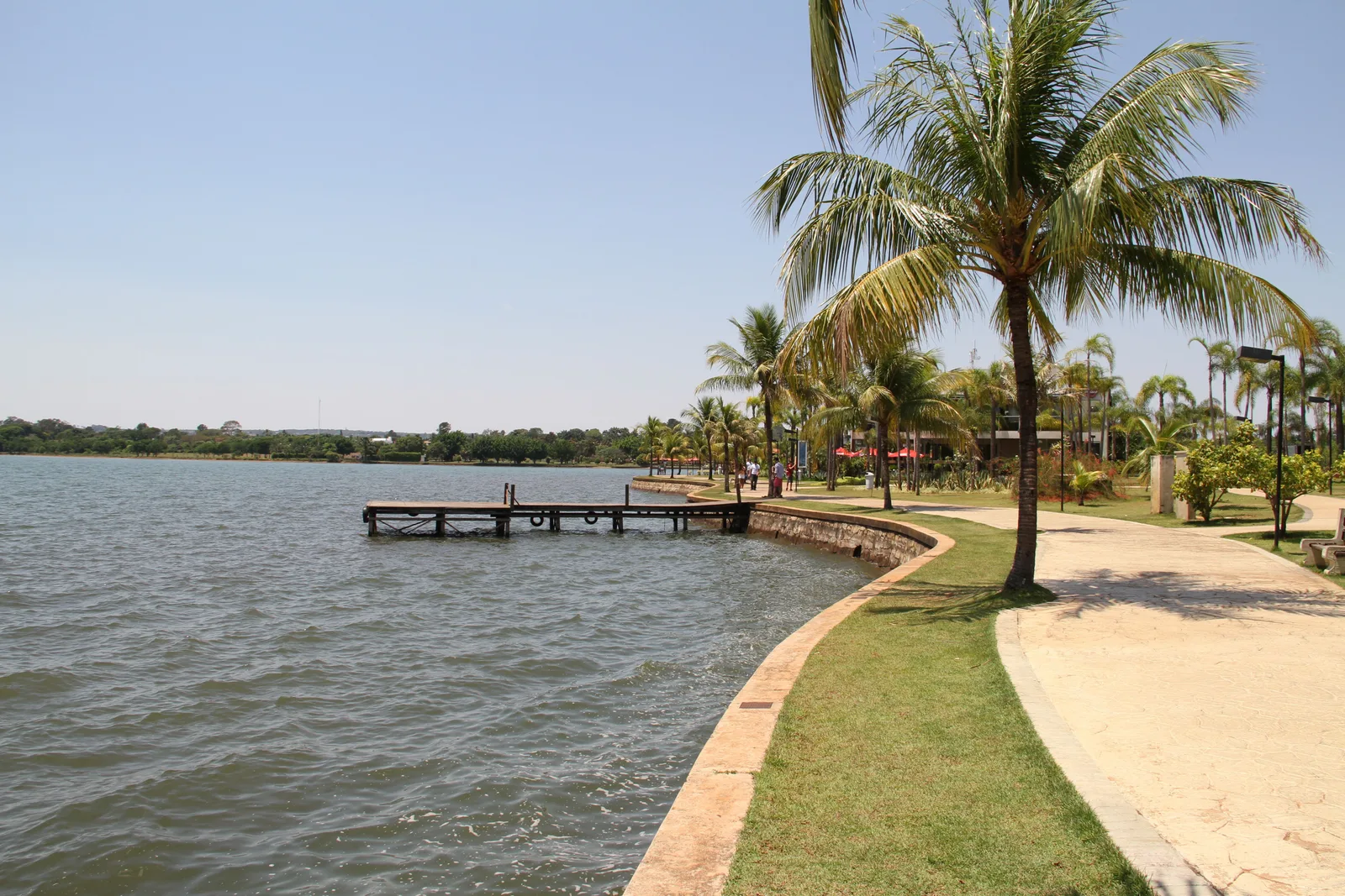 The waterfront at the Pontão do Lago Sul: a paved path curving along the lake past palm trees and a small pier, the water calm under a clear sky