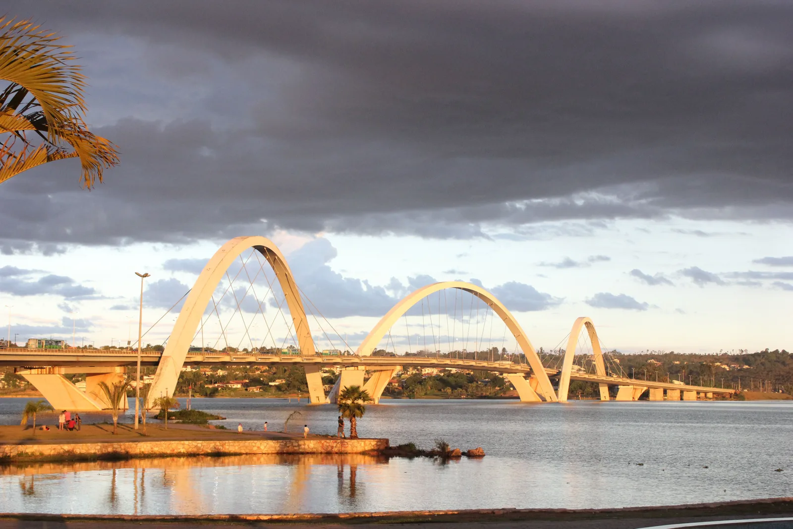 The Ponte JK at sunset, its three arches glowing gold under a dark cloudy sky and reflected in the calm water of Lago Paranoá
