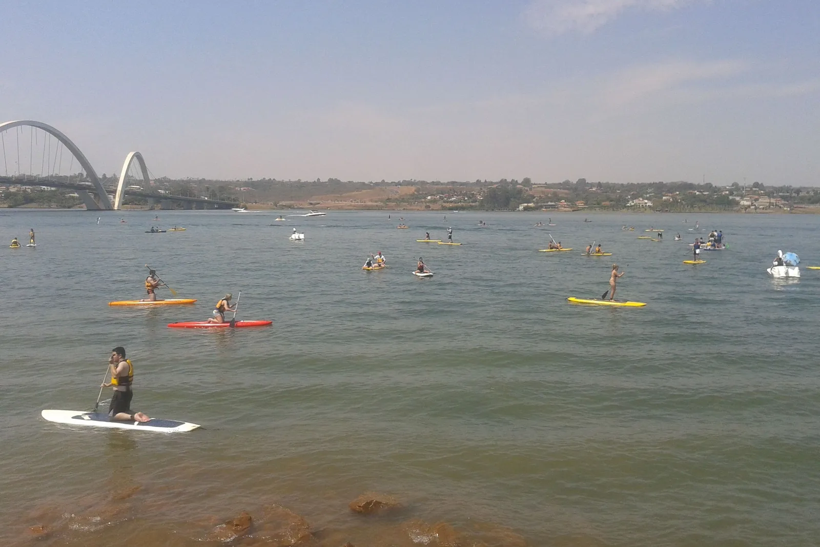 Stand-up paddlers and kayakers on Lago Paranoá on a hazy day, a man in a life vest kneeling on his board in the foreground and the arches of the Ponte JK on the horizon