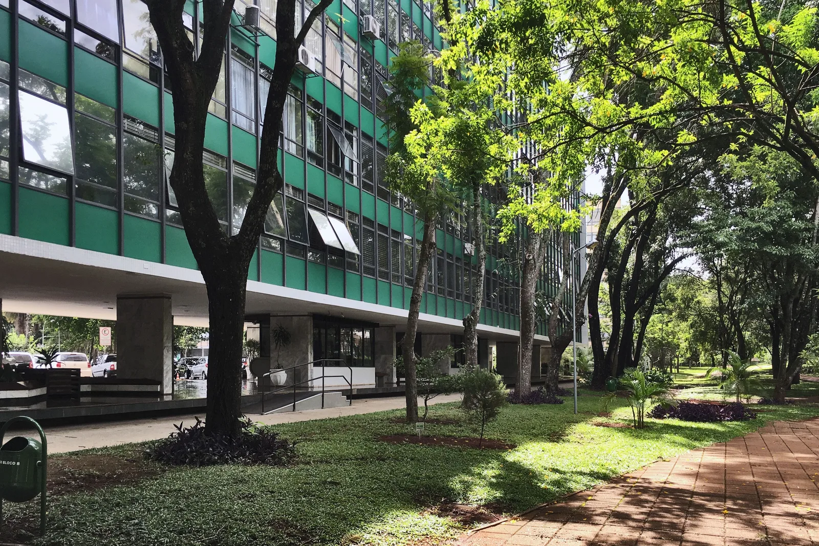 A superquadra in Asa Sul: a long apartment block with green-tinted windows raised on pilotis behind tall trees, a red brick path and lawns running past it