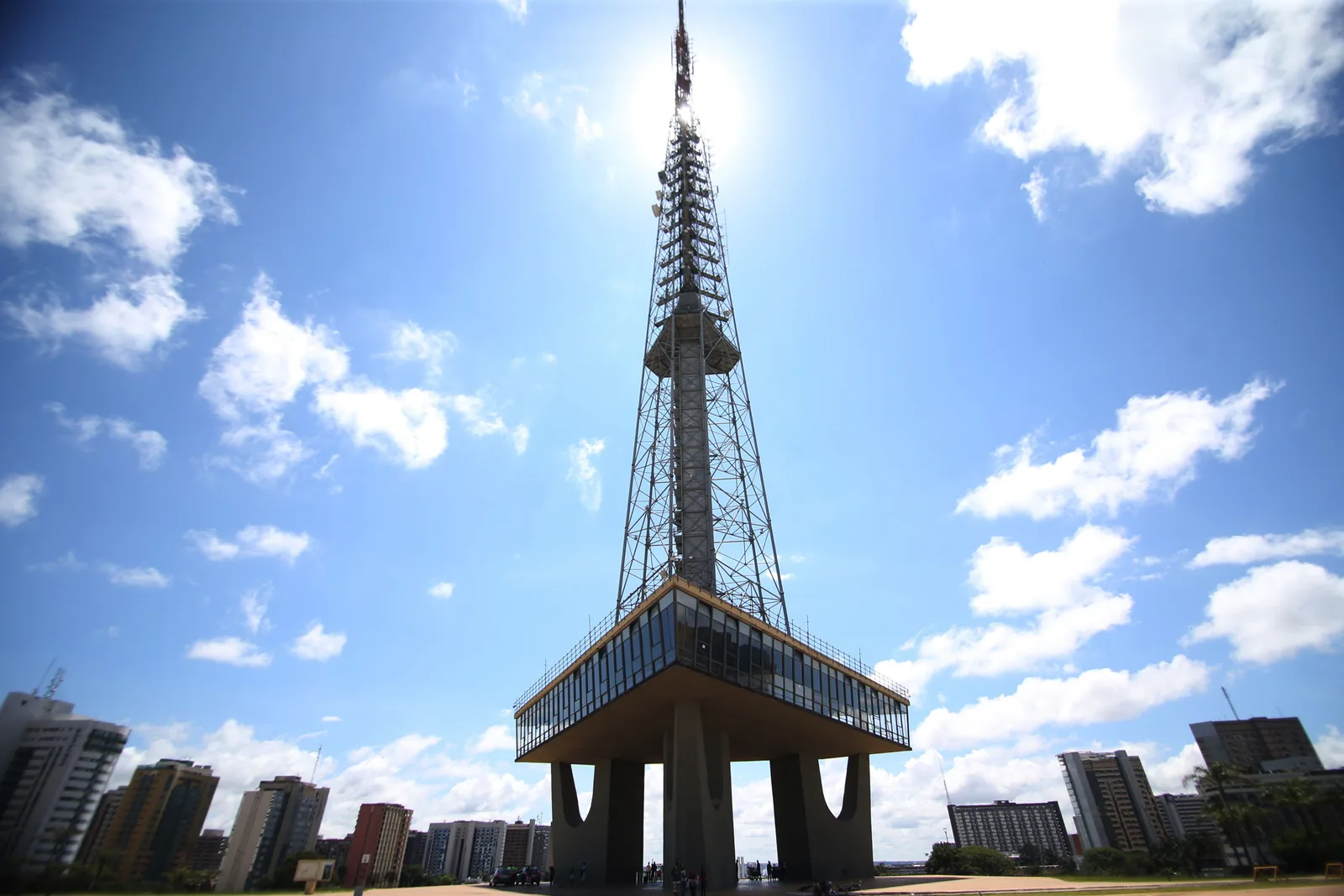 The Torre de TV from below: a tall steel lattice mast rising from a concrete platform into a bright blue sky with white clouds, apartment blocks low on the horizon
