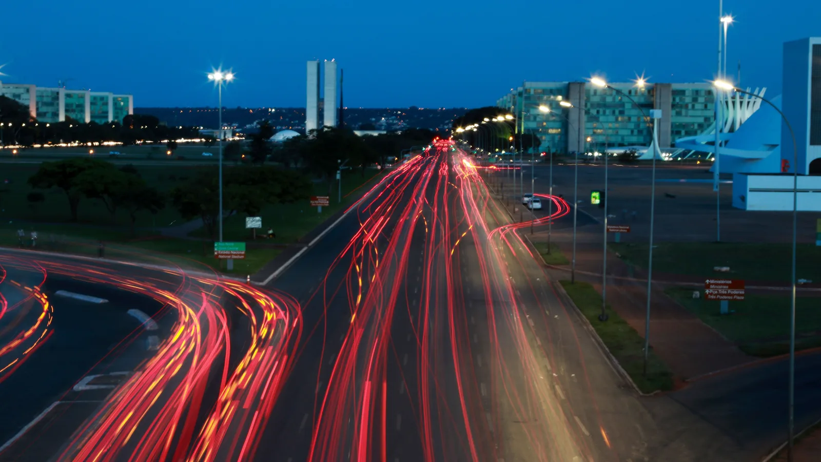 Brasília at dusk: red and white light trails of traffic sweeping along the Eixo Monumental toward the lit towers of Congress and the ministry blocks under a deep blue sky