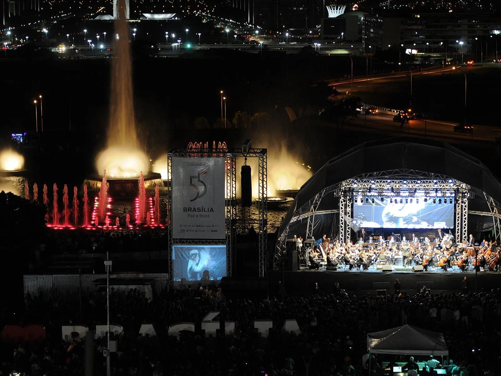 An orchestra on a brightly lit open-air stage at night at the Torre de TV during Brasília’s birthday celebrations, a crowd in front and the lit fountain at the left