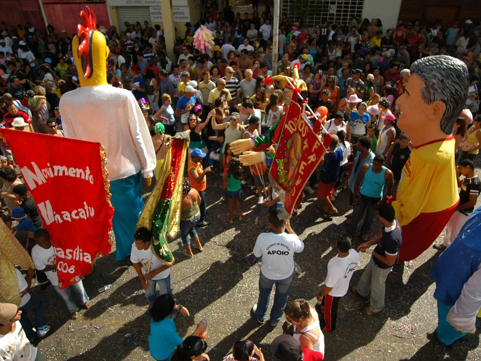 A Carnaval street bloco in Brasília: a dense crowd in costume around giant carnival puppets and a red banner on a sunny afternoon
