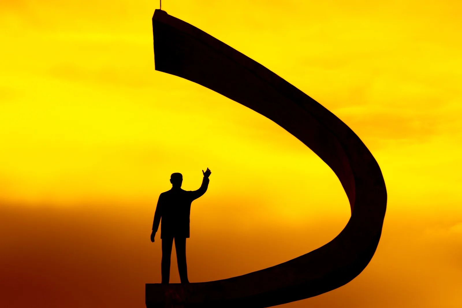 The statue of Juscelino Kubitschek on top of the Memorial JK, his arm raised, silhouetted inside the curved concrete crook of the tower against a vivid orange sky