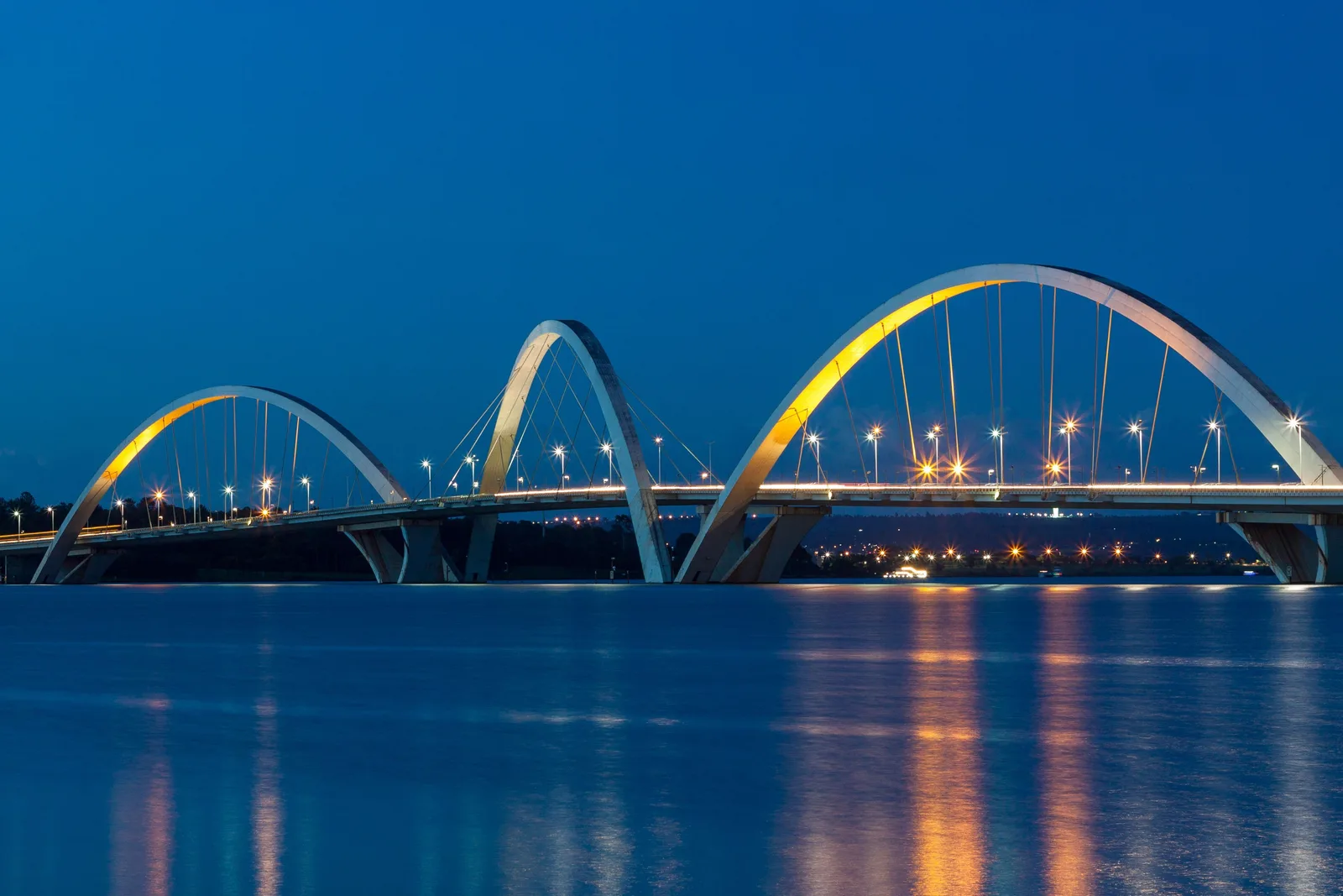The Ponte JK at blue hour, its three white steel arches lit from below and reflected in the dark water of Lago Paranoá