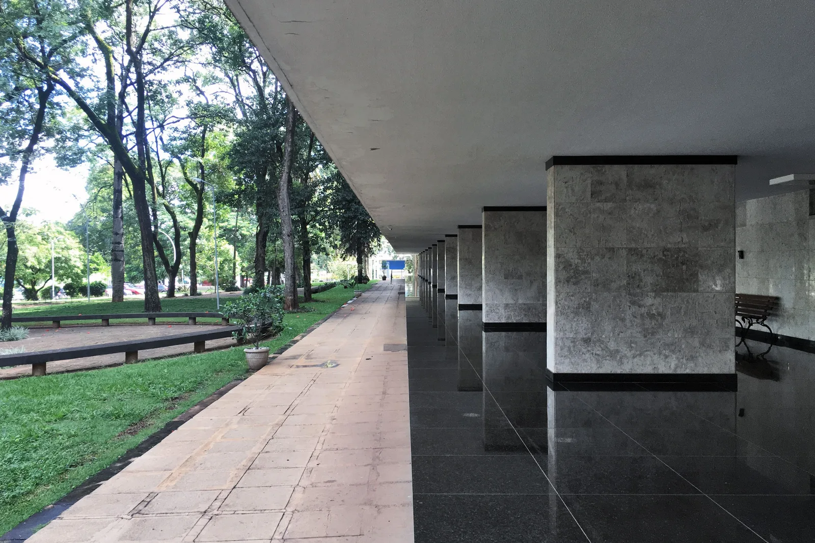 The open ground floor of a superquadra apartment block in Brasília, a paved walkway beside the marble-clad pillars, a lawn and trees on the left