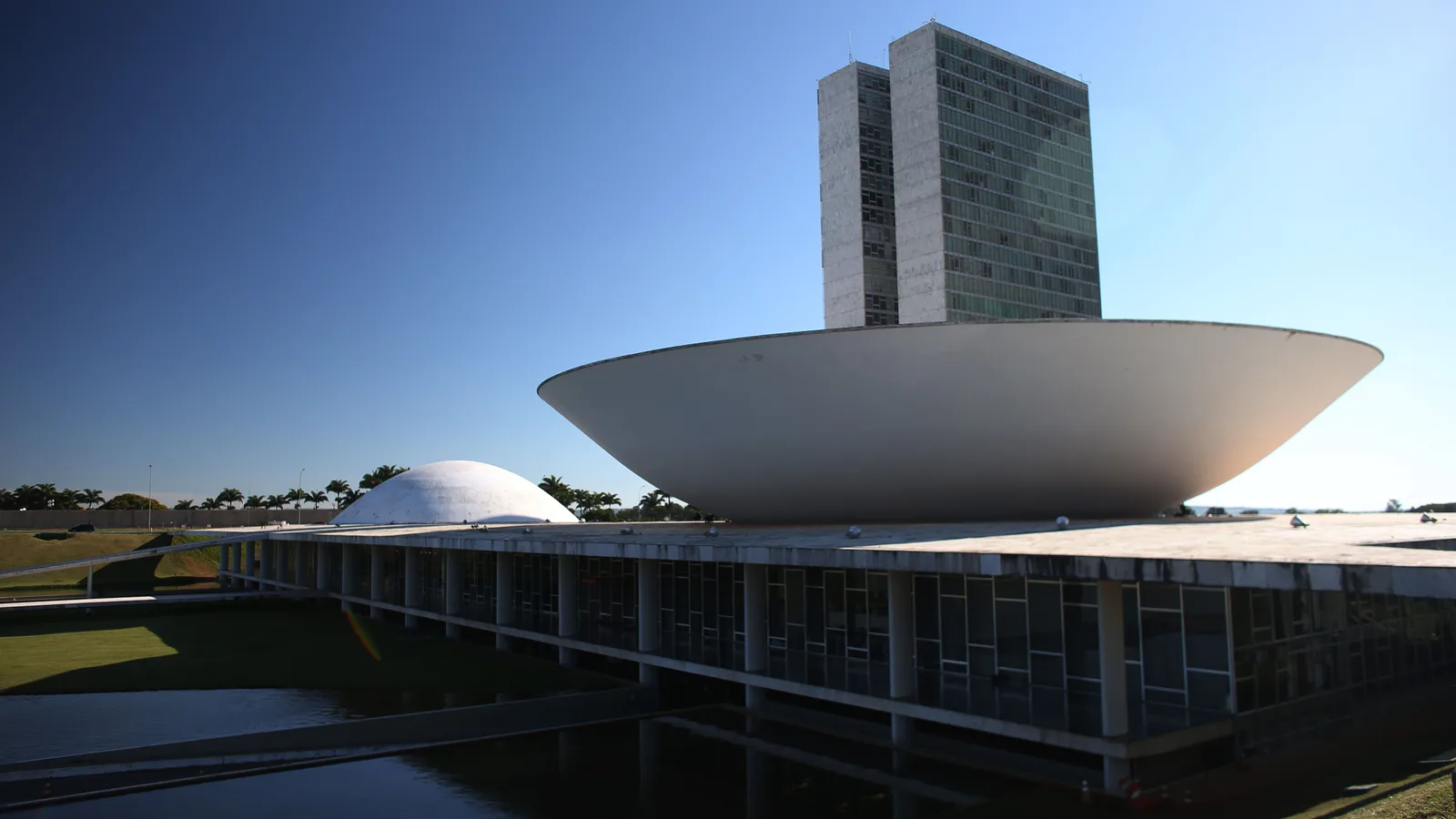 The National Congress in Brasília under a deep blue sky: the twin towers rising behind the shallow upturned bowl of the Chamber of Deputies, the low dome of the Senate at the left, and the long glazed base of the building above a dark reflecting pool