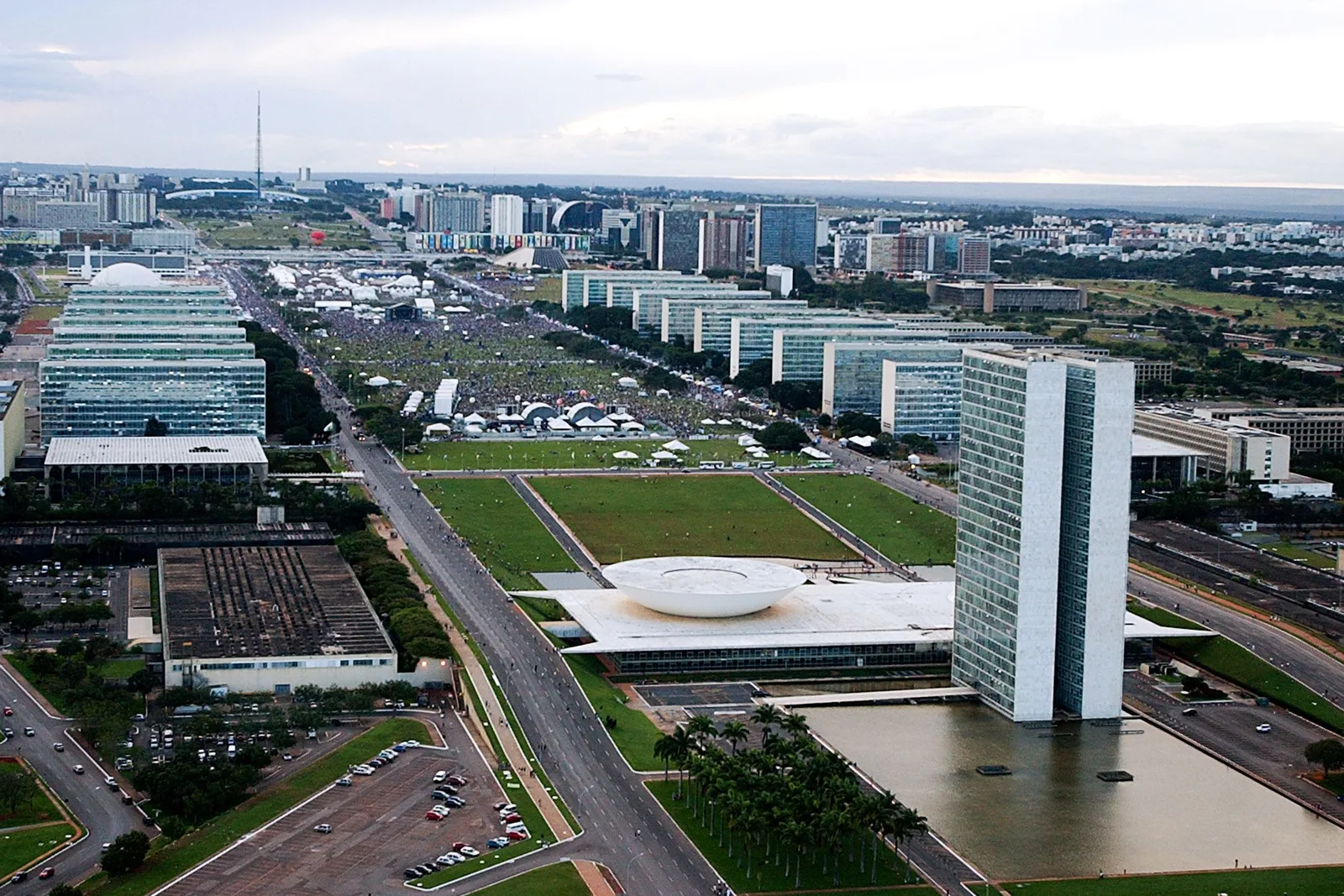 Congress from above, its twin towers and the bowl of the Chamber in the foreground, and the Esplanada dos Ministérios beyond packed with a crowd and white tents for a public event, the TV tower on the horizon