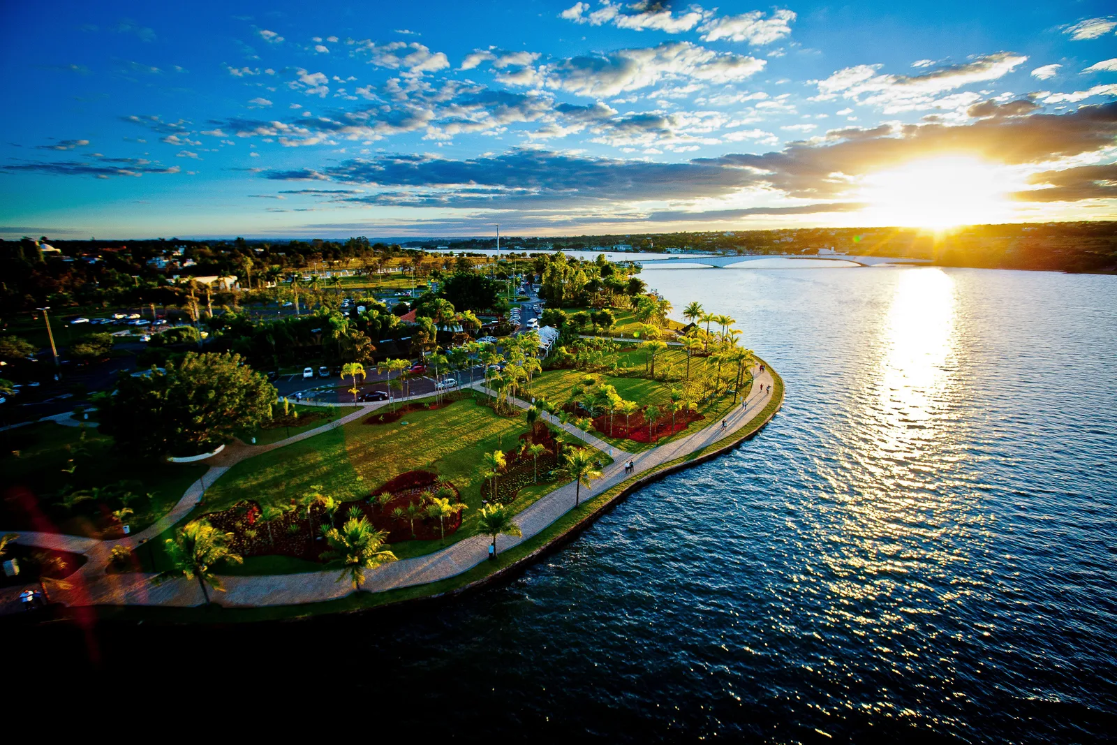 The sun setting over Lago Paranoá from the air, a wooded point of the shore with paths and lawns in the foreground and the light glittering on the water