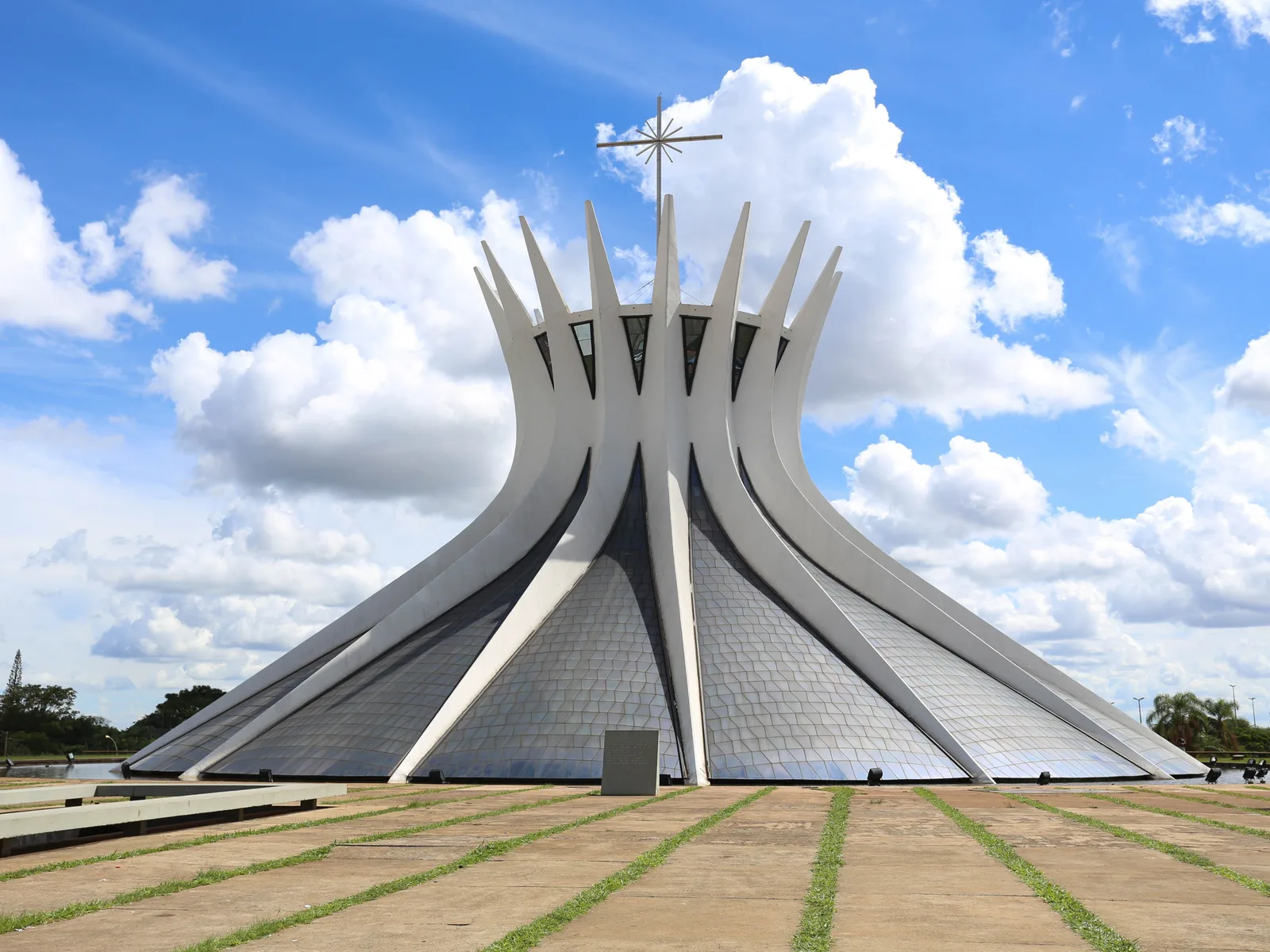The Cathedral of Brasília from the plaza: a ring of white curved concrete ribs gathering into a crown topped by a cross, the dark stained glass between them, under a blue sky piled with white cloud