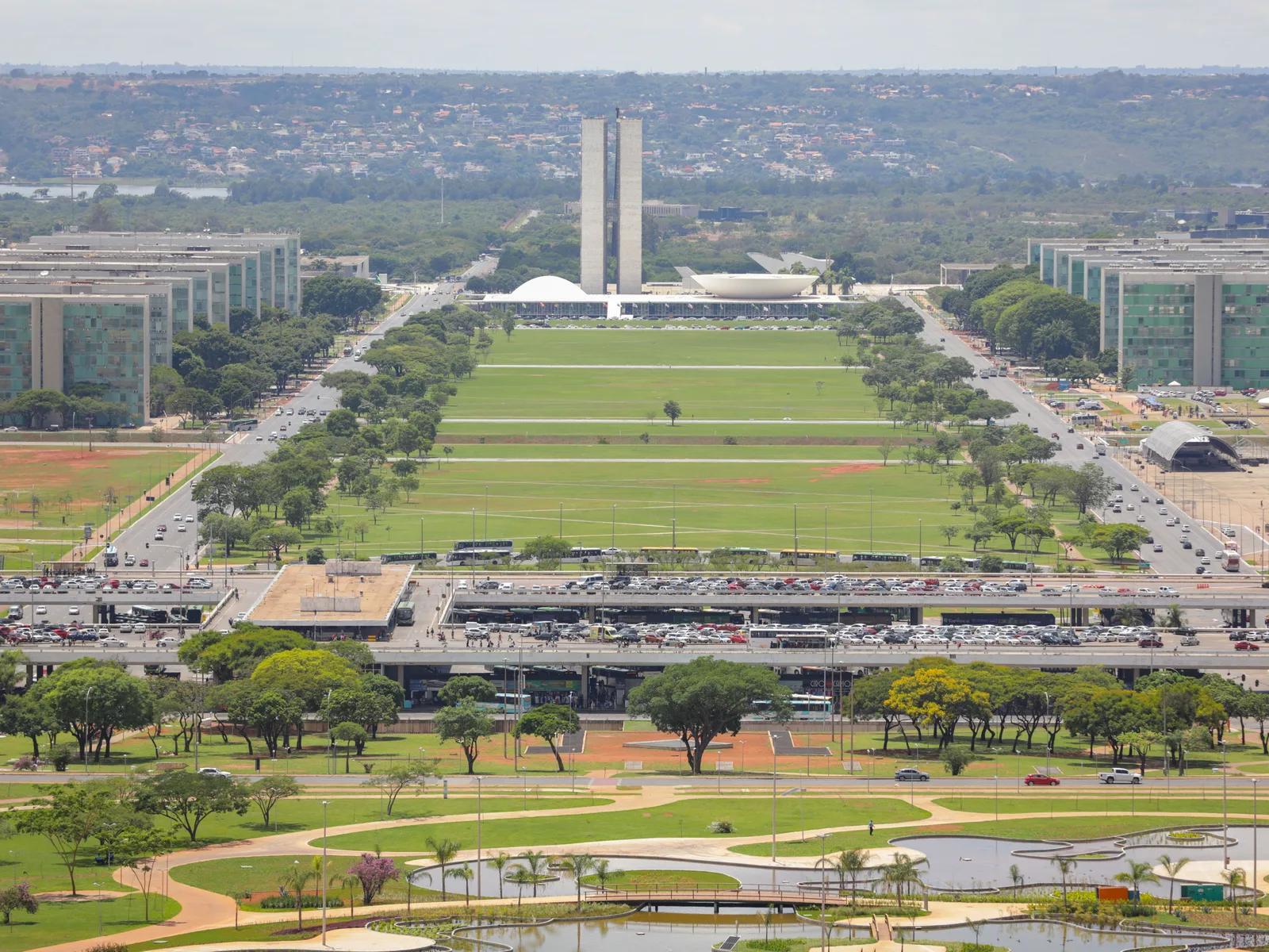 The Eixo Monumental from the Torre de TV: a wide green lawn running between two rows of ministry blocks toward the twin towers of Congress, the bus station platform and lines of trees in the foreground and the lake on the horizon