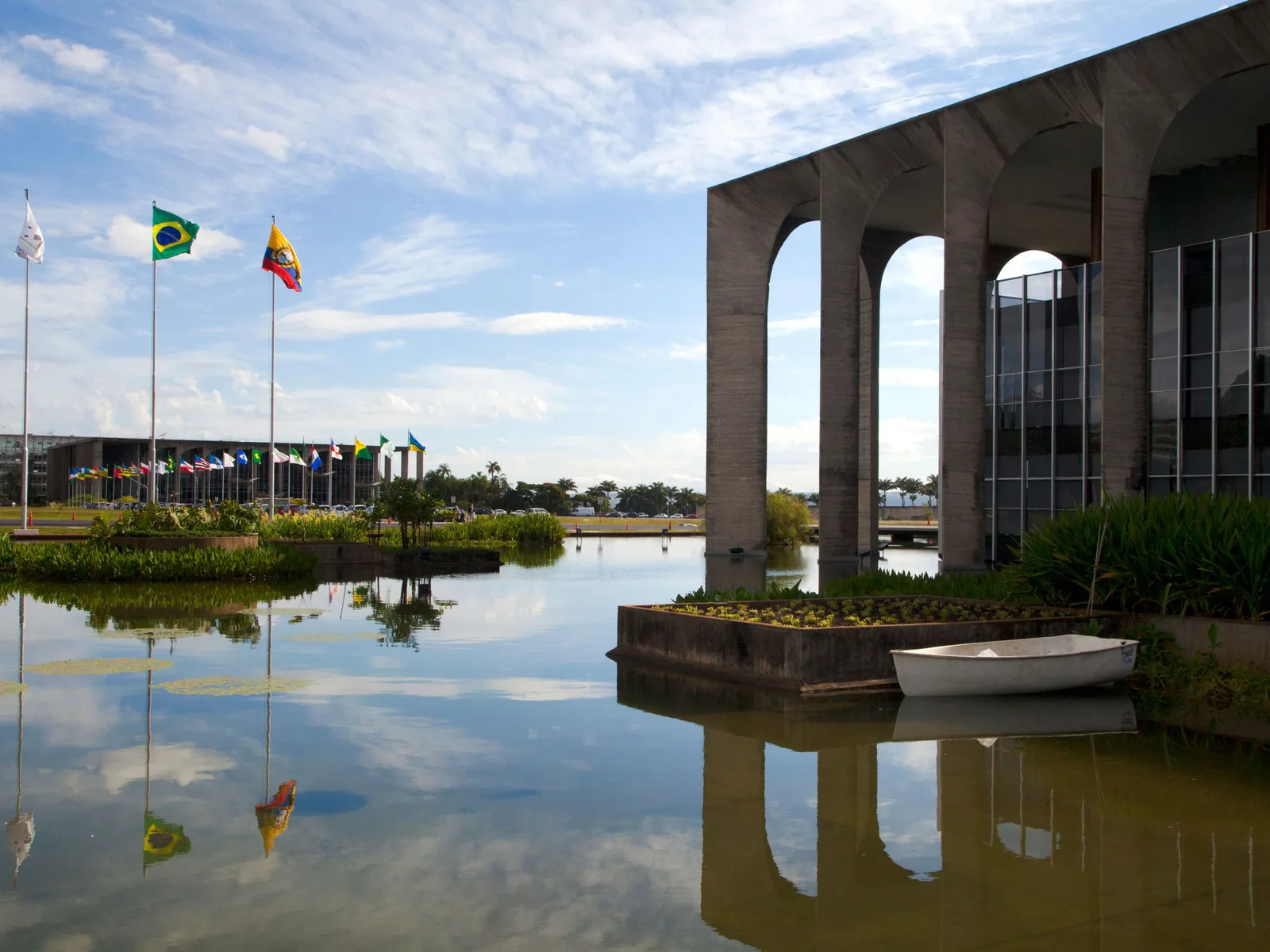 The Itamaraty Palace seen across its reflecting pool: tall slender concrete arches mirrored in the still water, tropical plants on islands in the pool and flags on tall poles beyond