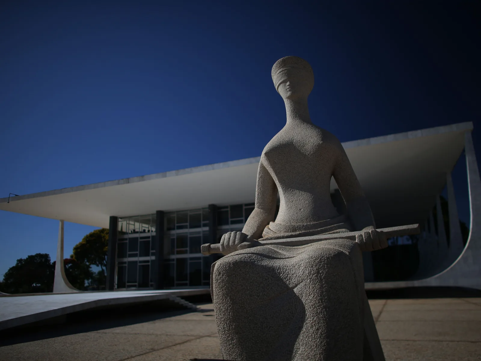 Alfredo Ceschiatti’s granite statue A Justiça, a seated blindfolded woman with a sword across her lap, in front of the low colonnade of the Supreme Court in the Praça dos Três Poderes under a deep blue sky