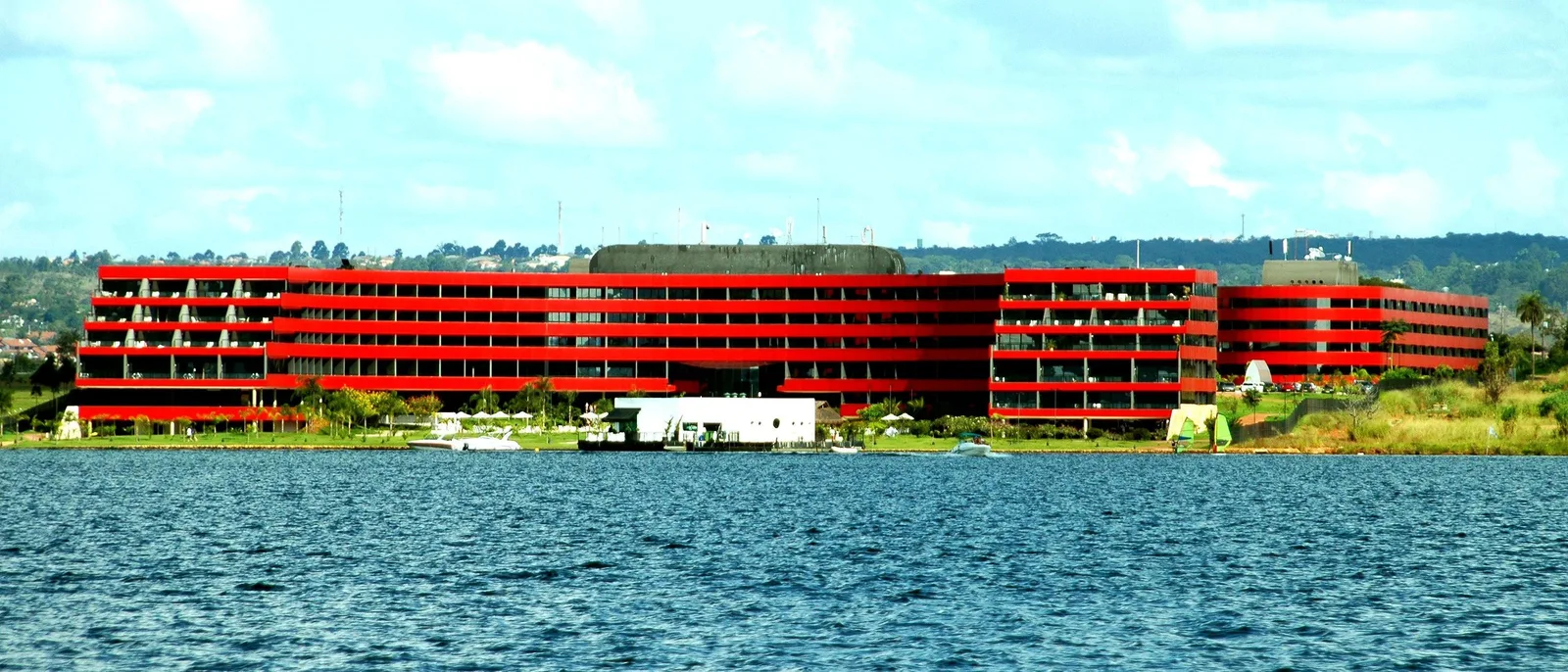 A long lakeside hotel painted bright red, its terraced floors running along the shore of Lago Paranoá, a small white building on a jetty and moored boats at the water’s edge, and choppy blue water in the foreground