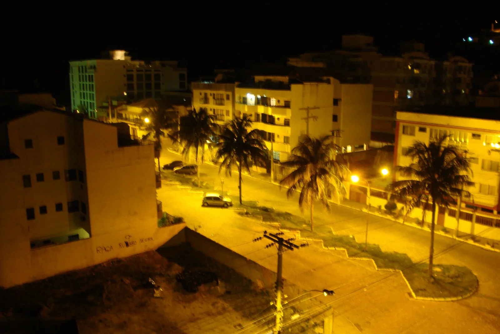 A street in Arraial do Cabo at night seen from above under orange streetlights: four- and five-story apartment buildings, a row of palm trees along the road and a few parked cars
